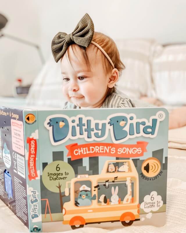 A baby in a large green bow headband sits on a bed, looking at the Ditty-Bird AUS "Children's Songs" USB-C rechargeable musical book, featuring colorful animal illustrations and playing classic children's songs.