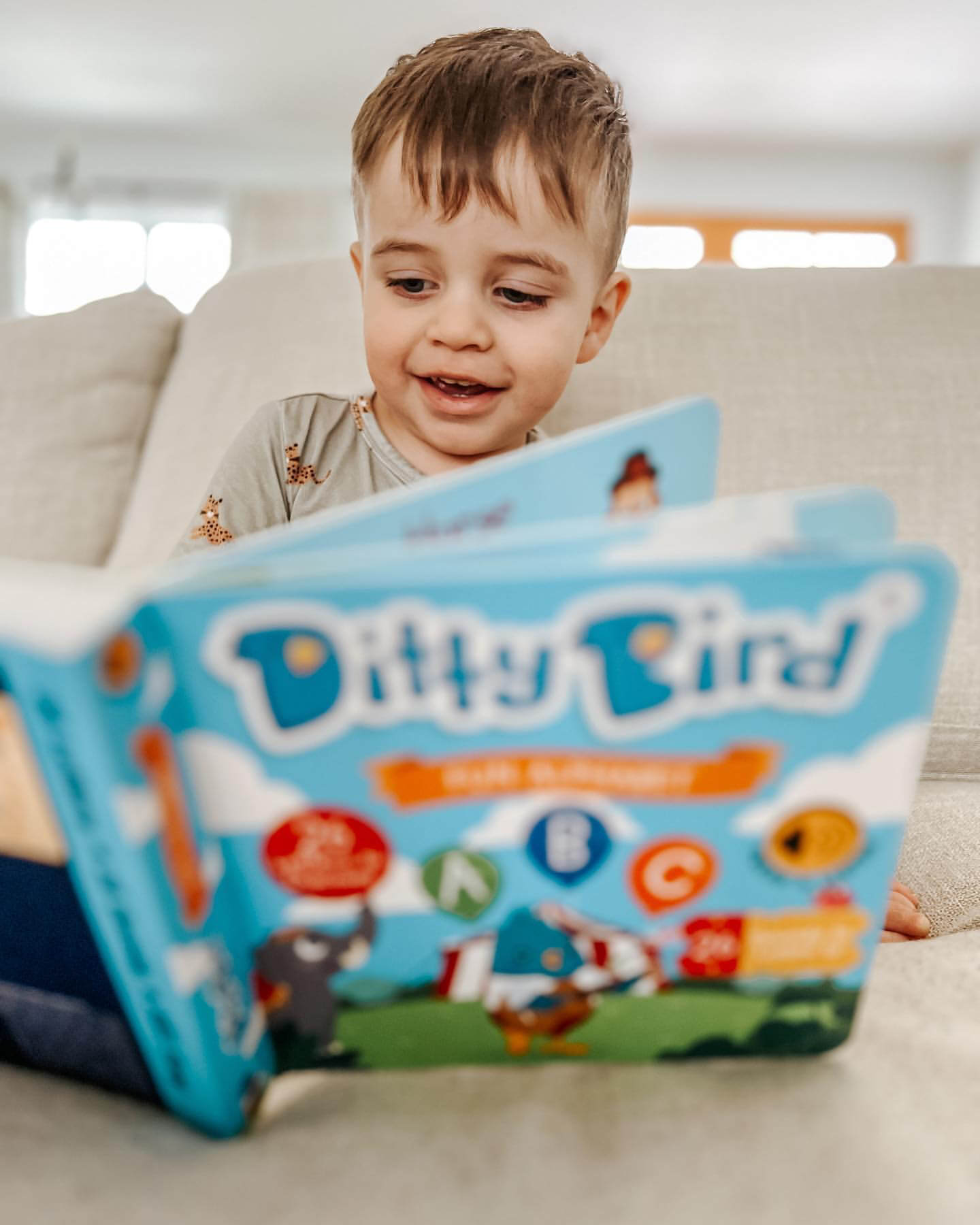 A young boy smiles on the couch as he explores the Ditty-Bird AUS ABC Fun Alphabet book, USB-C rechargeable, featuring playful illustrations and bright colors to encourage early literacy skills.