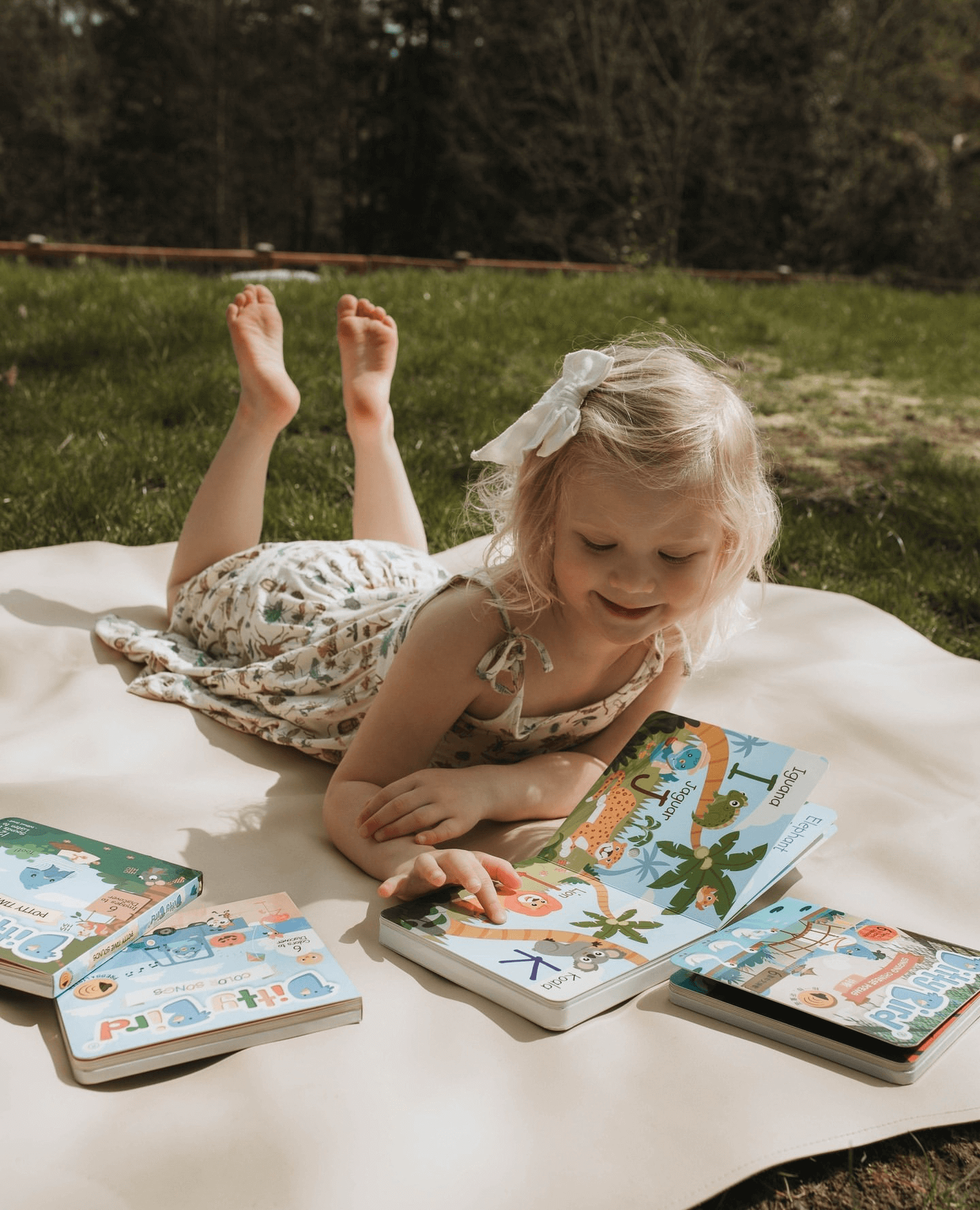 A young girl with blonde hair and a white bow smiles as she reads the Ditty-Bird AUS ABC Fun Alphabet, a USB-C rechargeable interactive book, building early literacy skills while lying on a blanket outdoors.