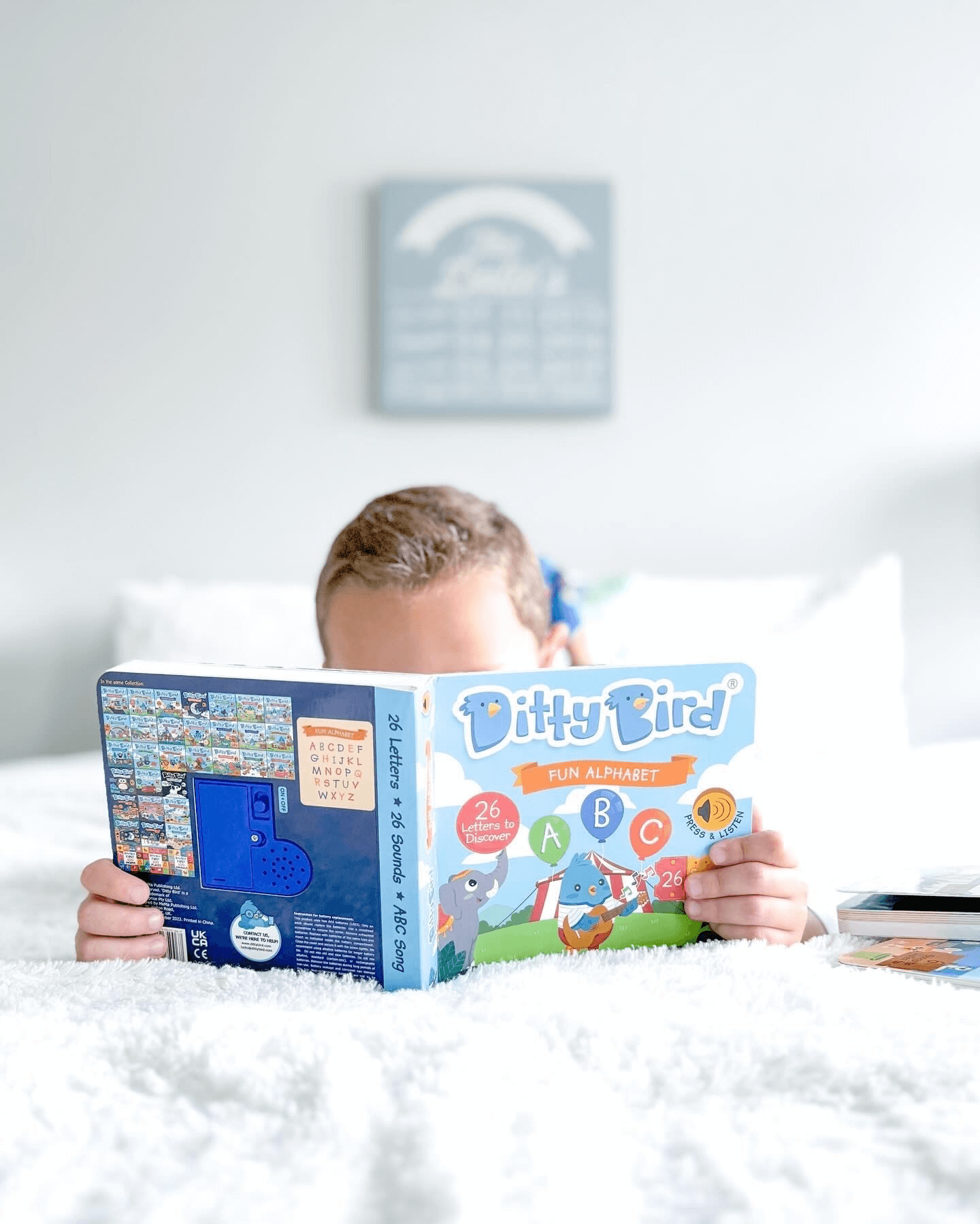 A child lies on a white bed reading the Ditty-Bird AUS ABC Fun Alphabet book (USB-C rechargeable), with just the top of their head visible above the open book. A blurred wall art piece hangs in the background, emphasizing early literacy skills.