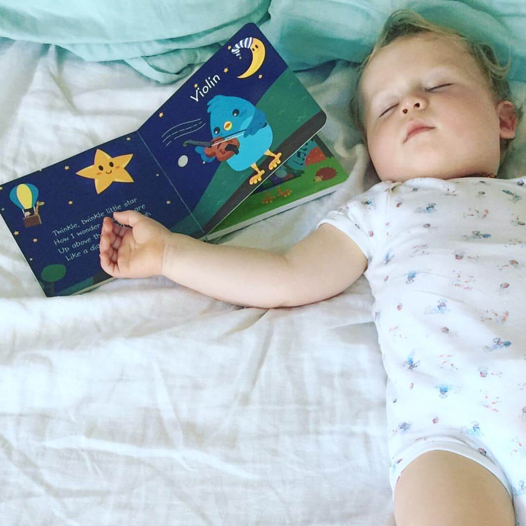 A sleeping toddler in a white onesie lies on a bed with one arm outstretched, touching an open Ditty-Bird AUS Children's Songs book (USB-C rechargeable), featuring colorful illustrations of a star, moon, and a blue bird playing a violin.