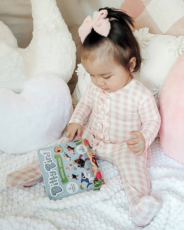 A toddler in pink plaid pajamas sits on a white knitted blanket, holding an open Ditty-Bird AUS Animal Songs interactive musical book (USB-C rechargeable), surrounded by plush pillows.