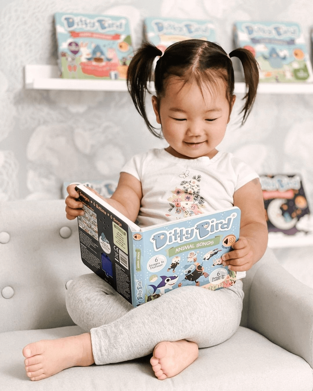 A young child with pigtails, wearing a white shirt and gray leggings, sits cross-legged on a chair and smiles while enjoying Ditty-Bird AUS’s USB-C rechargeable interactive musical book, Animal Songs. Colorful books line the shelf in the background.