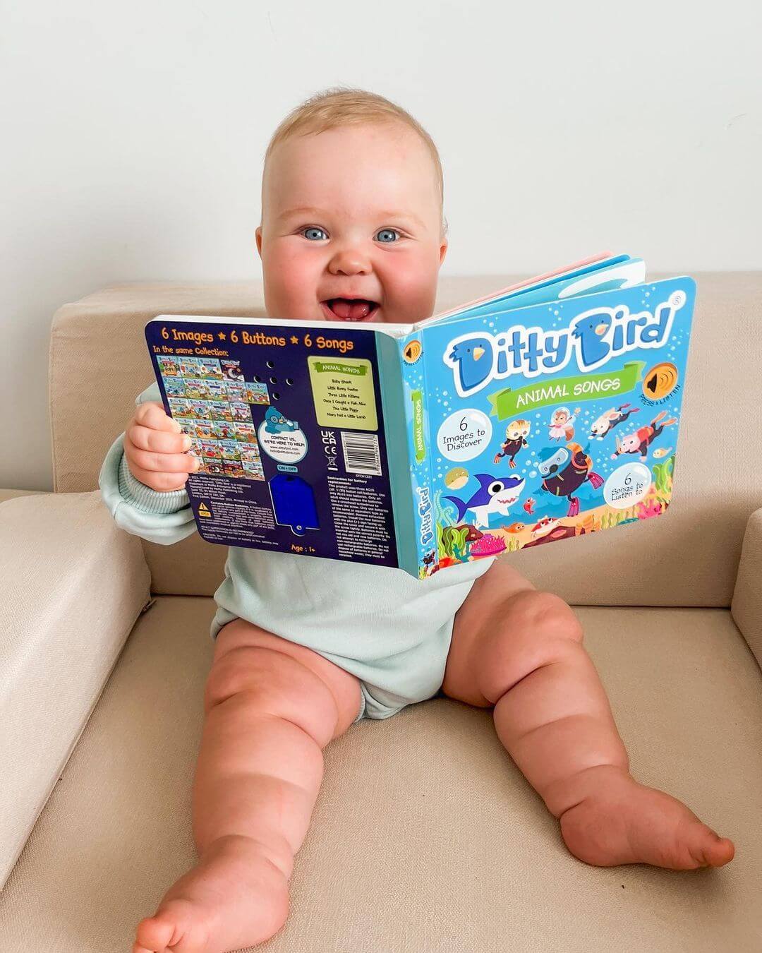 A smiling baby in a light blue outfit sits on a beige couch, happily exploring the Ditty-Bird AUS Animal Songs | USB-C rechargeable interactive musical book for kids.