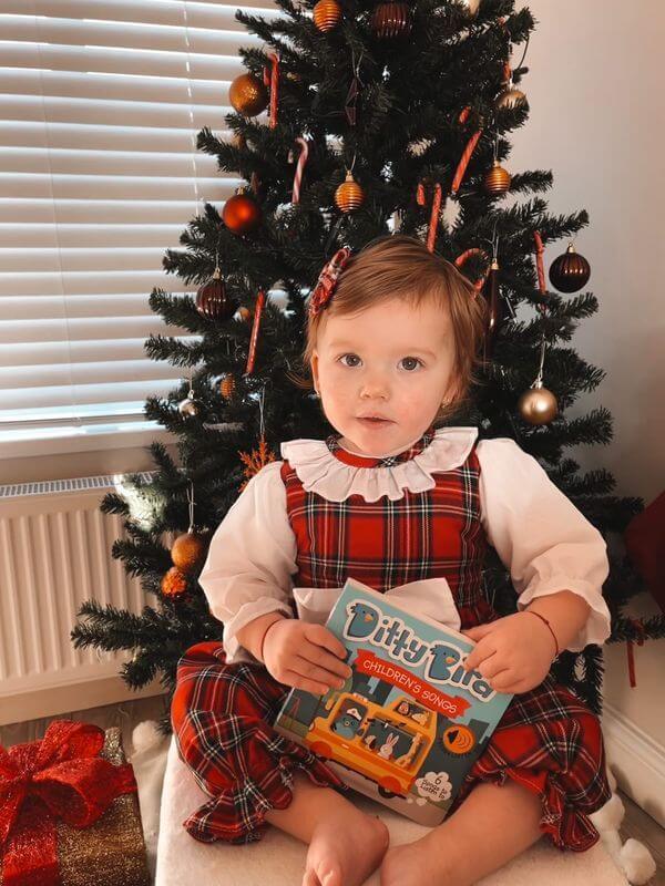 A young child in a red plaid dress sits by a decorated Christmas tree, holding the Ditty-Bird AUS Children's Songs | USB-C rechargeable book. Presents are visible under the tree.