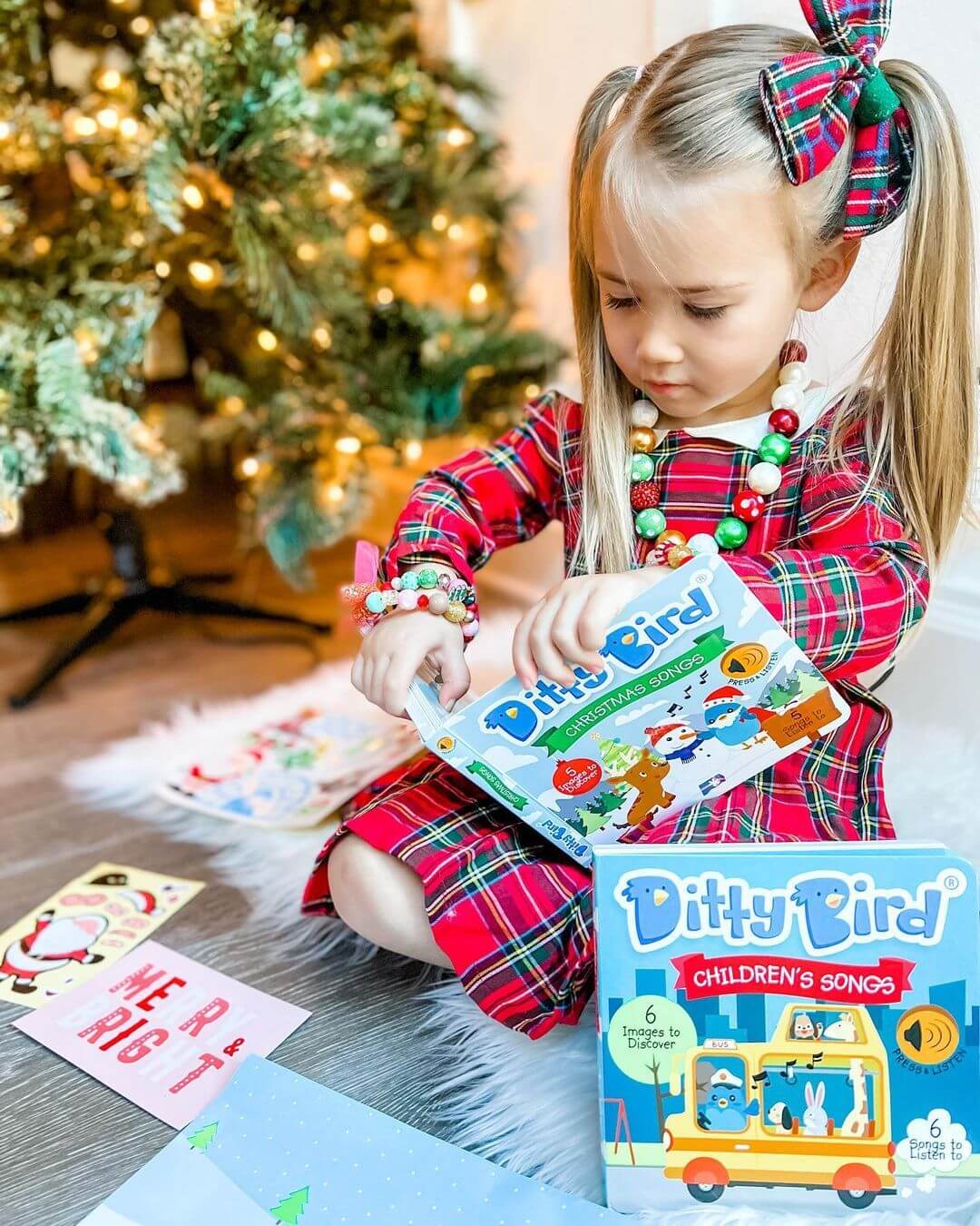 A young girl in a red plaid dress opens Ditty-Bird AUS’s USB-C rechargeable "Children's Songs" interactive sound book by the Christmas tree, surrounded by gift wrap and cards as she discovers fun melodies.