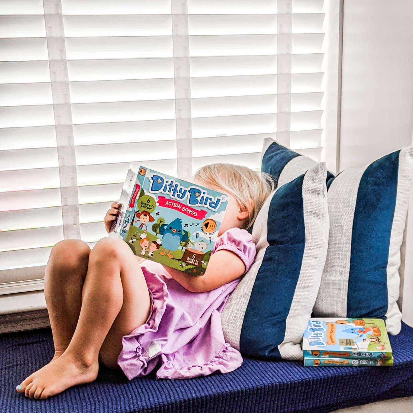 A young child in a pink dress sits on a window seat with blue pillows, holding and looking at a Ditty-Bird AUS children’s music book. Another Ditty-Bird AUS Action Songs | USB-C rechargeable sound book rests beside her. White blinds cover the window behind.