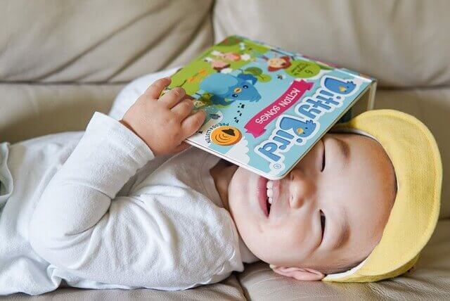 A cheerful baby in a yellow cap lies on a couch, playfully holding the Ditty-Bird AUS "Action Songs" children's music book (USB-C rechargeable) over their face.