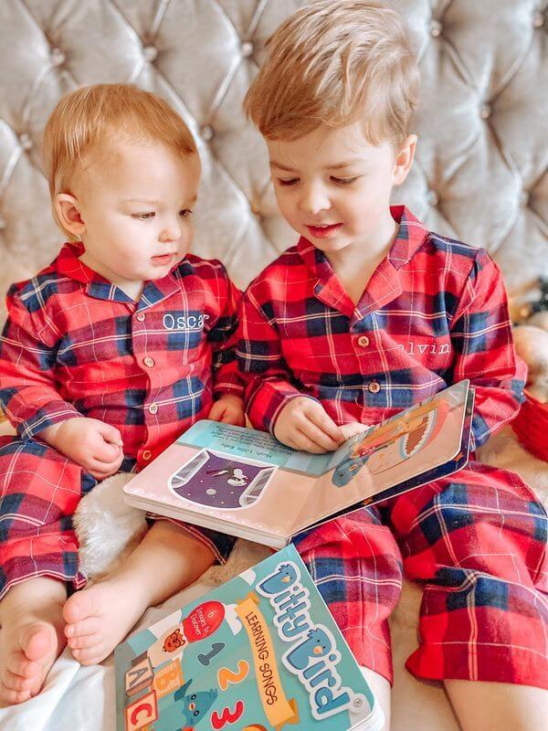 Two young children in matching red plaid pajamas sit together on a bed, looking at picture books—a cozy scene that would make the perfect memory or inspire a thoughtful first birthday gift. The older child reads aloud while the younger one attentively watches.