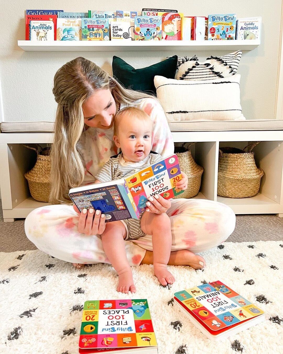 A woman sits on the floor with a baby on her lap as they read the Ditty-Bird AUS 100 Words Sound Book (USB-C rechargeable). Two more books lie open nearby, with colorful children's books displayed on a shelf in the background.