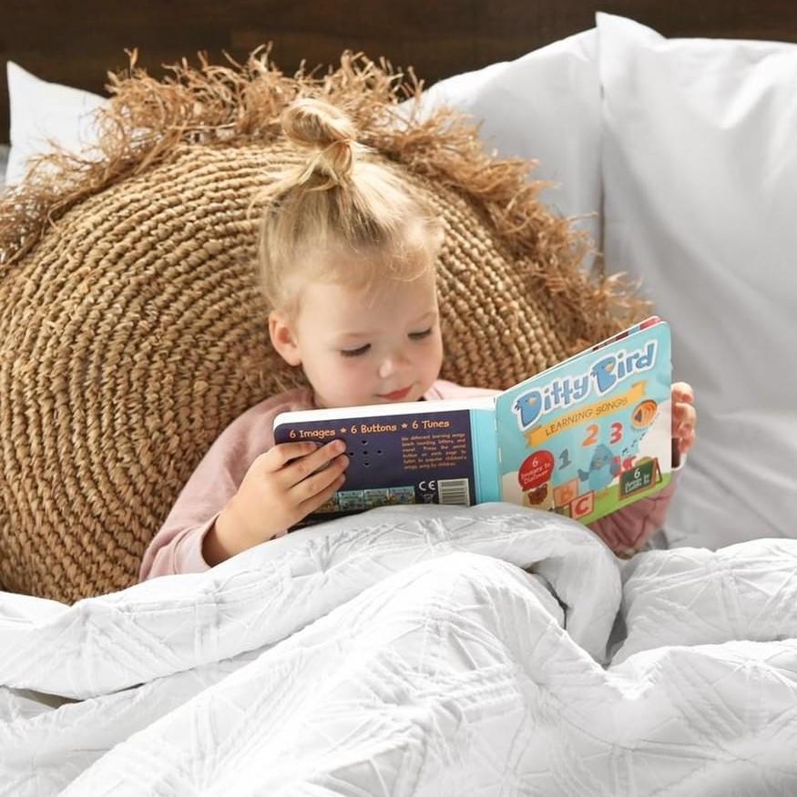 A young blonde child sits in bed, propped against a round woven pillow, reading the Ditty-Bird AUS "Learning Songs | USB-C rechargeable" interactive sound book, wrapped in a cozy white quilt.