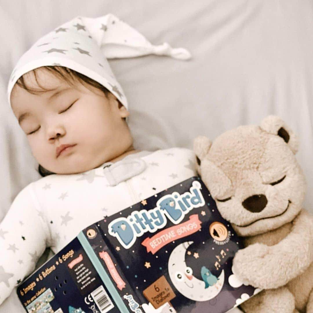 A baby wearing a star-patterned hat and pajamas sleeps while cuddling a beige teddy bear and holding a Ditty Bird Bedtime Songs book, an ideal educational toy for babies 6-12 months, on a soft, light-colored surface.