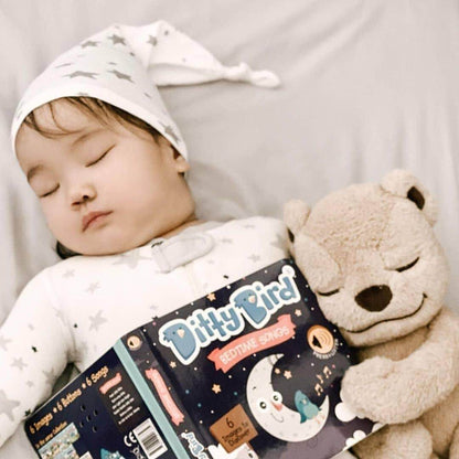 A baby wearing a star-patterned hat and pajamas sleeps while cuddling a beige teddy bear and holding a Ditty Bird Bedtime Songs book, an ideal educational toy for babies 6-12 months, on a soft, light-colored surface.